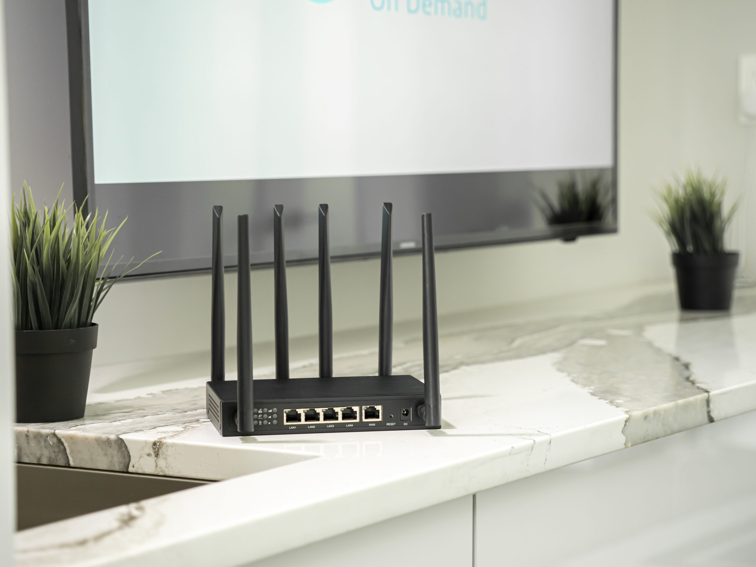 X-LINK router displayed on a white surface with decorative plants and office monitor in background.
